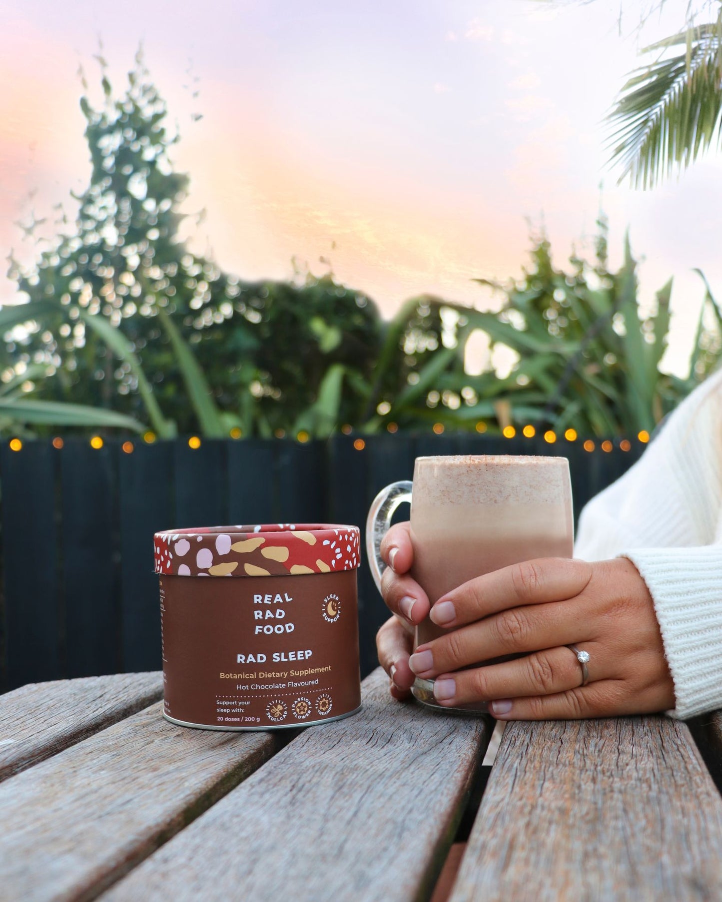 Hands holding a mug of Real Rad Food Rad Sleep sleepy hot chocolate beside a Rad Sleep botanical supplement tub during a calming sunset nighttime ritual